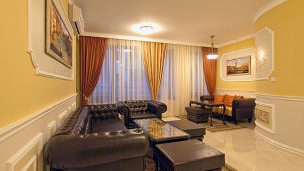 A living room with a sectional sofa, a small table, and a chair. There is a window with curtains, a painting on the wall, and a chandelier. - Luxury Apartment Venice - 1 (Sofia)
