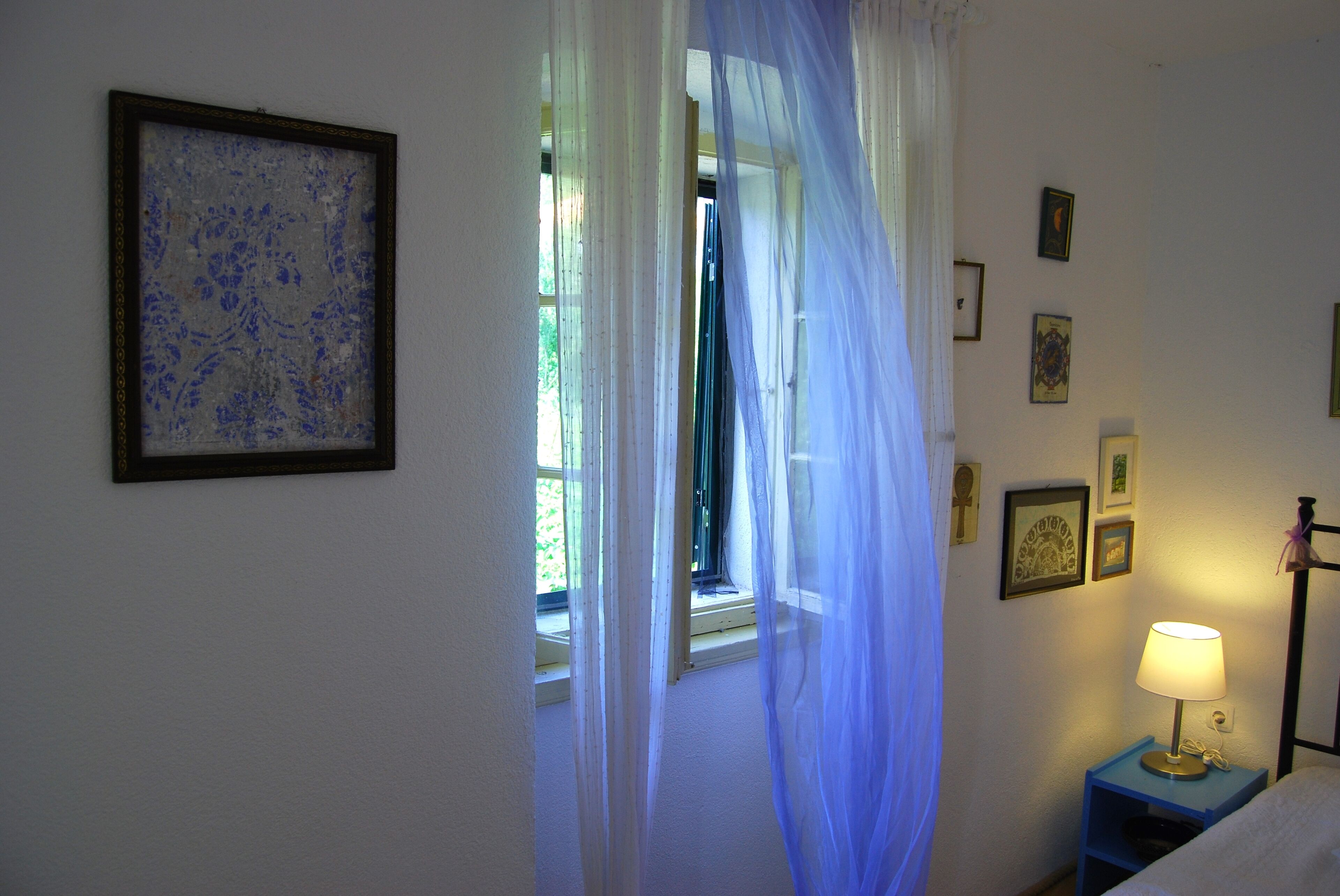 A bedroom with a bed, a nightstand with a lamp, a window with sheer curtains, and a wall with framed pictures.