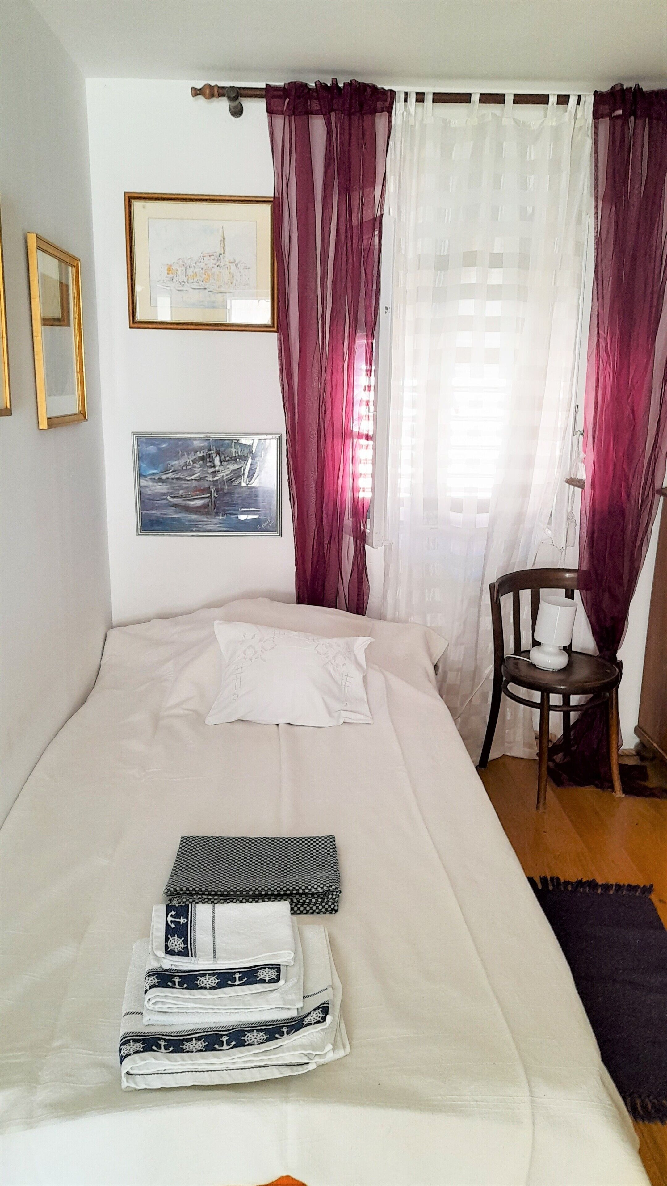 Una cama bien hecha con edredón y almohadones blancos, una pequeña silla de madera con una lámpara blanca, y dos cuadros en la pared.