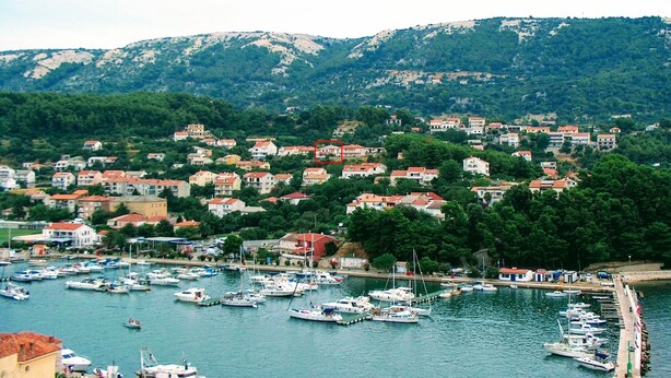 A coastal town with a marina filled with boats, surrounded by residential buildings and green hills in the background. - Apartment With View (Rab)