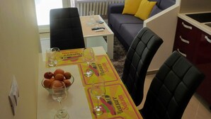 A dining area with a table set for two, a sofa with yellow cushions, and a view of the outside through large windows. - Apartment With View (Rab)