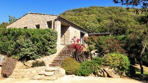 A stone building with a staircase leading to a covered entrance, surrounded by lush greenery and a hillside in the background. - Stone villa with heated pool close to the most beautiful beaches! (Figari)