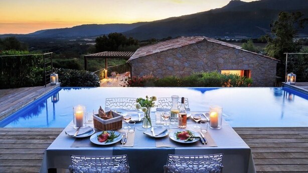 A dining table set for two with a view of a mountainous landscape and a stone building in the background. - Stone villa with heated pool close to the most beautiful beaches! (Figari)