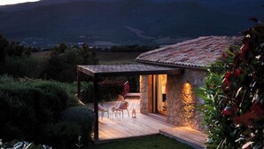 A modern house with a stone exterior and a wooden deck, surrounded by lush greenery and mountains in the background. - Stone villa with heated pool close to the most beautiful beaches! (Figari)
