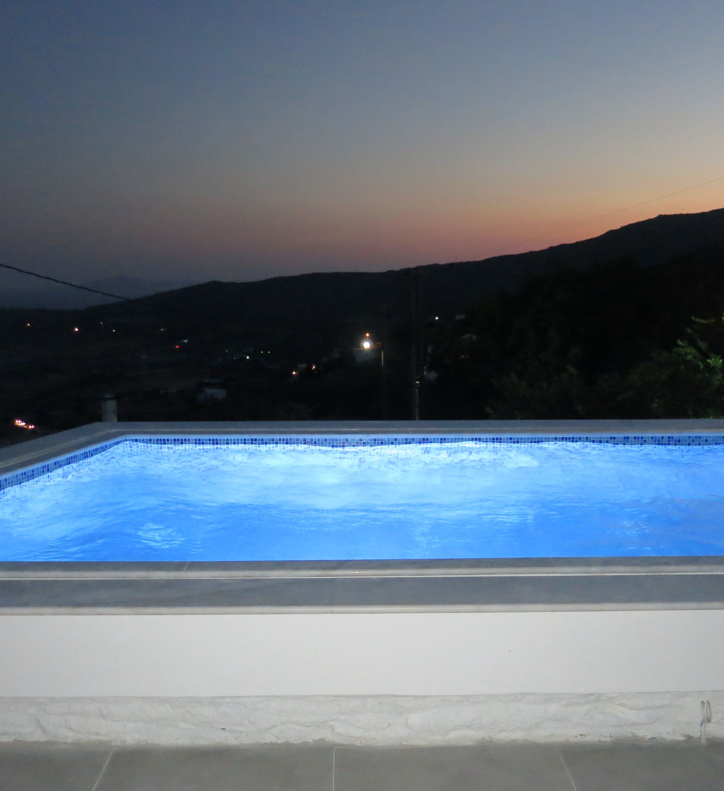 Una piscina al anochecer con vistas a colinas distantes y algunas luces de fondo.