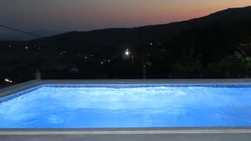 Piscina ao entardecer com vista para colinas distantes e algumas luzes ao fundo.