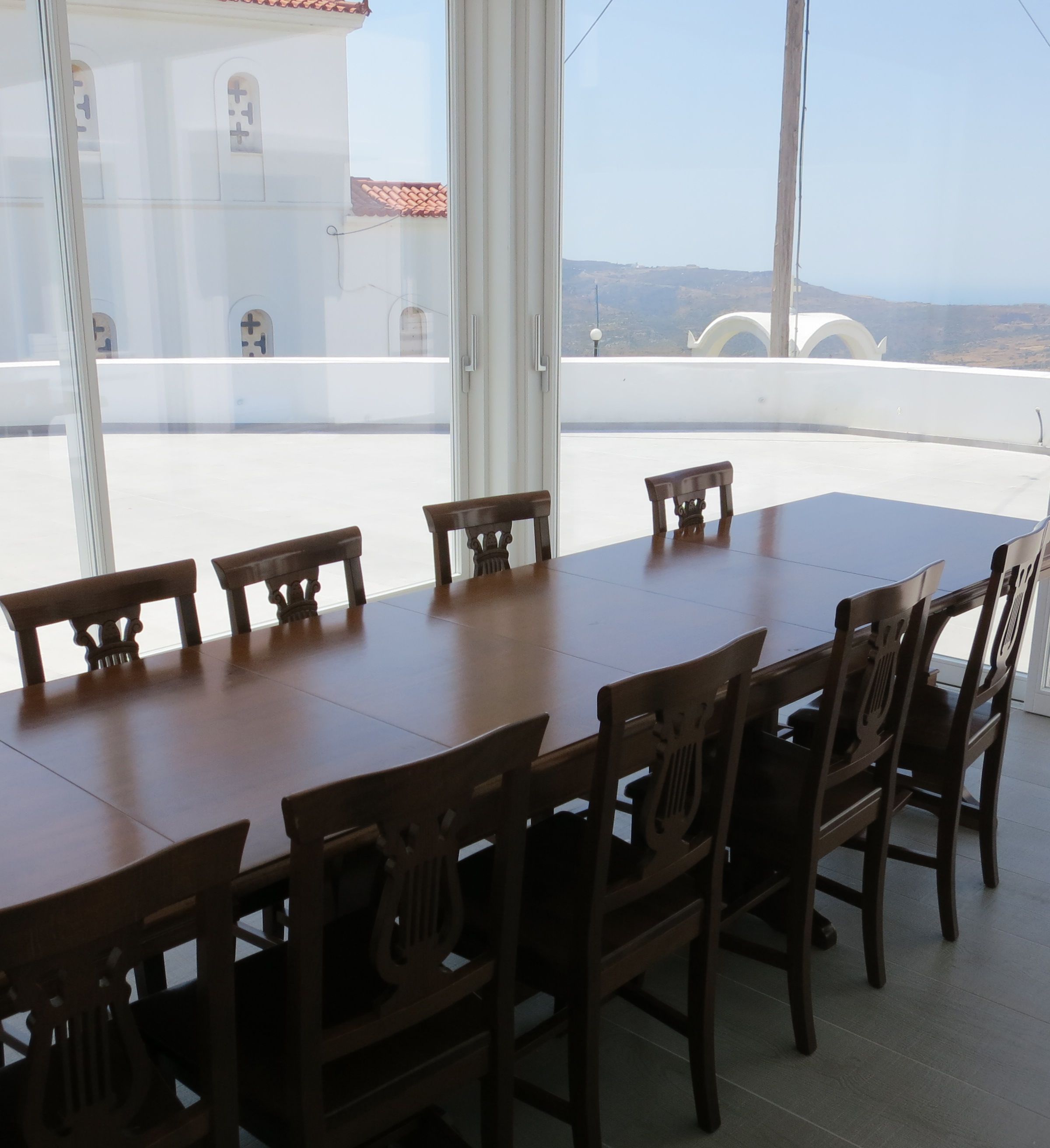 Uma sala de jantar com uma mesa de madeira comprida e cadeiras, grandes janelas com vista para edifícios e colinas.