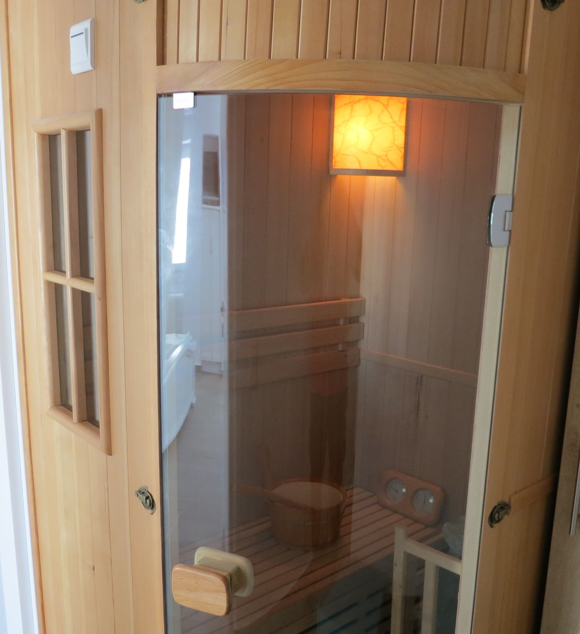 Uma sauna de madeira com porta de vidro, banco de madeira, mesa pequena e aquecedor fixado na parede.