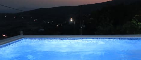 Una piscina al anochecer con vistas a colinas distantes y algunas luces de fondo.