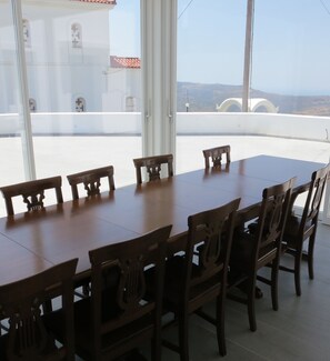 A dining area with a long wooden table and chairs, large windows with a view of buildings and hills. - Room-andoni's House in Pitrofos Andros i (Andros)
