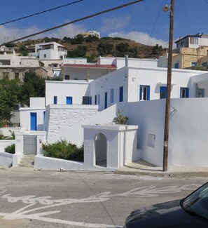 Un edificio blanco con puertas y ventanas azules, una calle pavimentada con carril bici, y un paisaje montañoso al fondo.