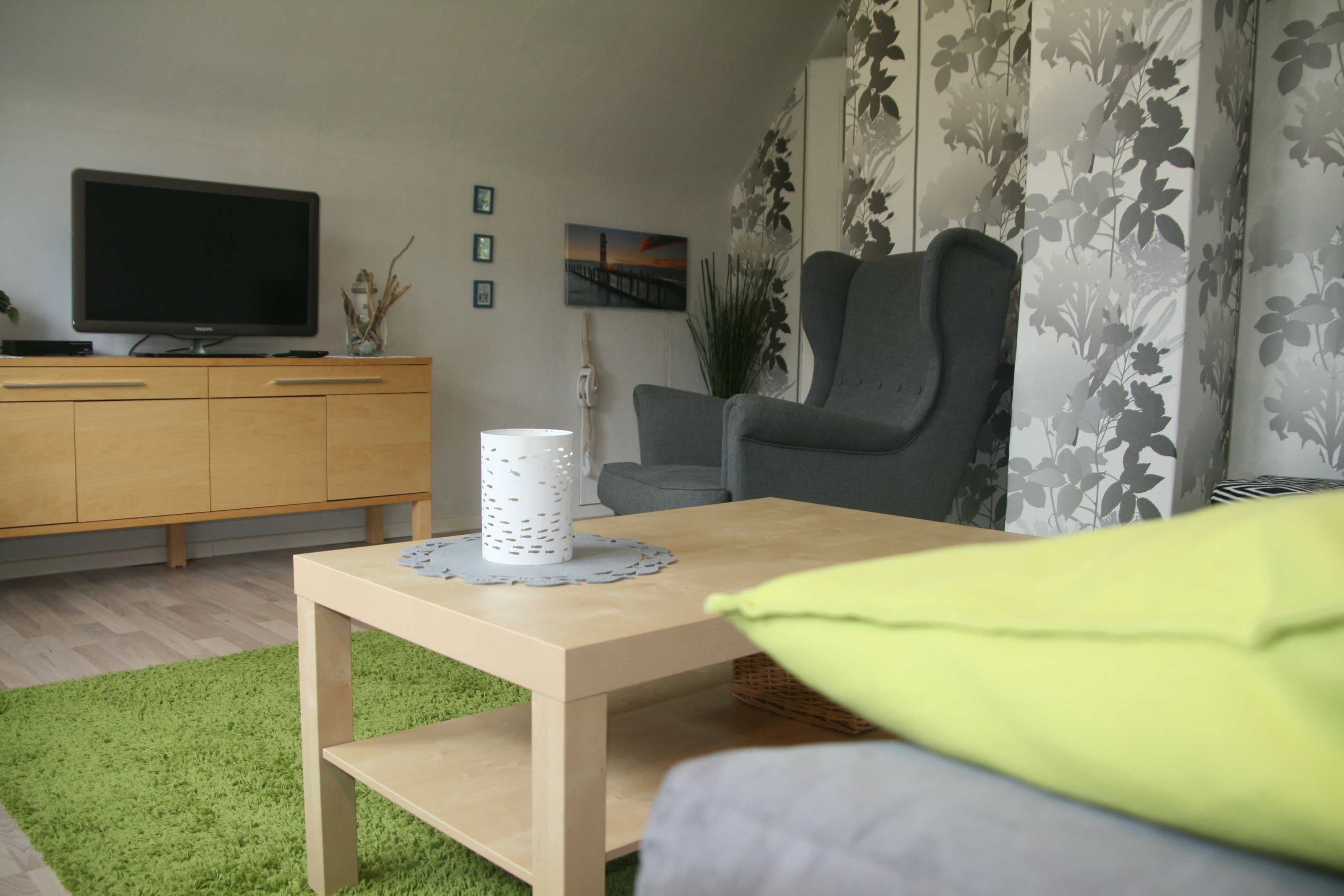 A living room with a television, a wooden cabinet, a grey armchair, a small table with a white candle, and a green rug.