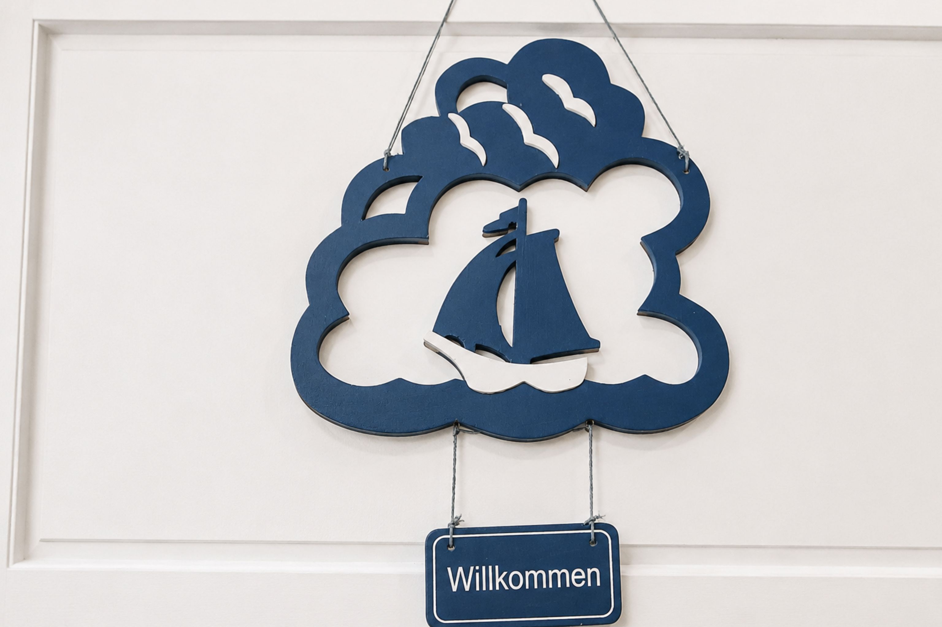 En dekorativ väggdekoration med en segelbåt och moln, och en skylt under som visar texten ”Willkommen”.