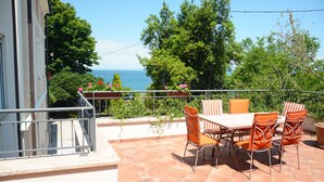 En udendørs terrasse med et bord og stole, med udsigt over vand og grøn vegetation.