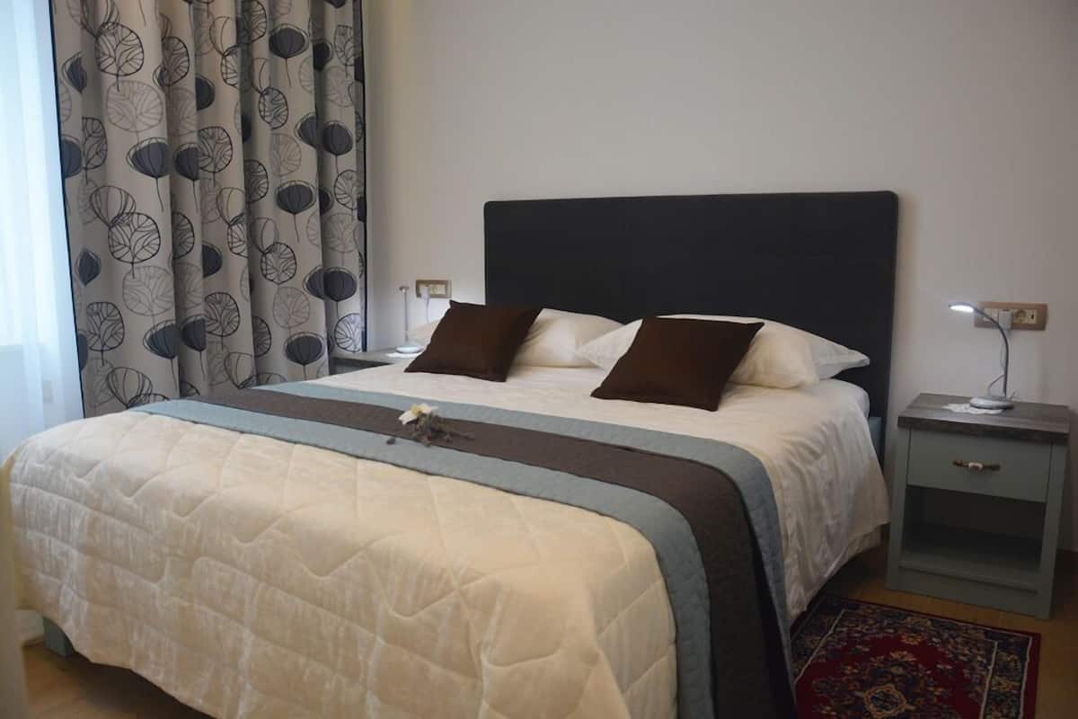 A neatly made bed with a dark headboard, a bedside table with a lamp, and a patterned curtain.
