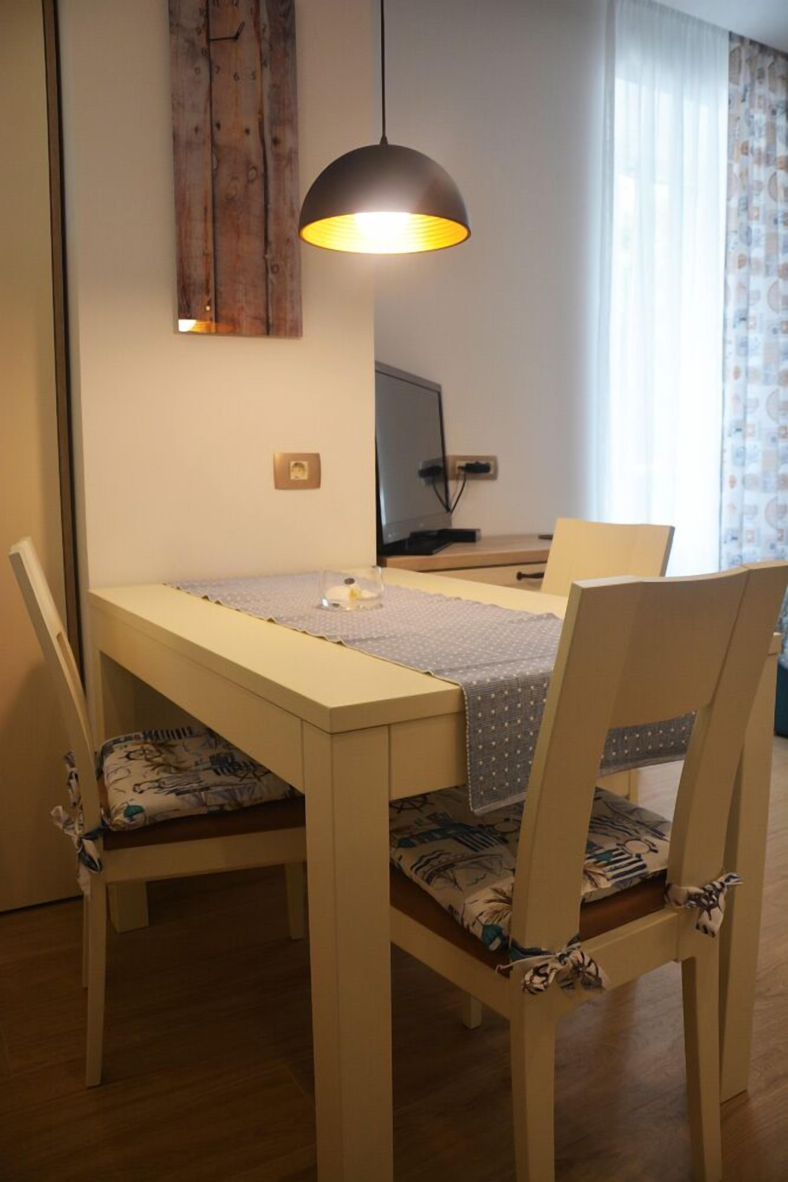 A dining area with a wooden table, four chairs, a pendant light, and a television on a stand.