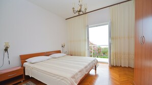 Quarto com cama de madeira, mesa de cabeceira, abajur, lustre e vista para uma varanda com cortinas.