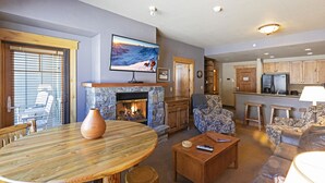 A cozy living room with a stone fireplace, a television mounted above it, and a dining area with a wooden table and chairs. - Top floor ski condo with direct ski in and out. Hiking and biking too! Hot tub (Whitefish)