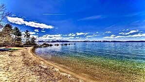 A clear beach with calm water, trees, and mountains in the background.