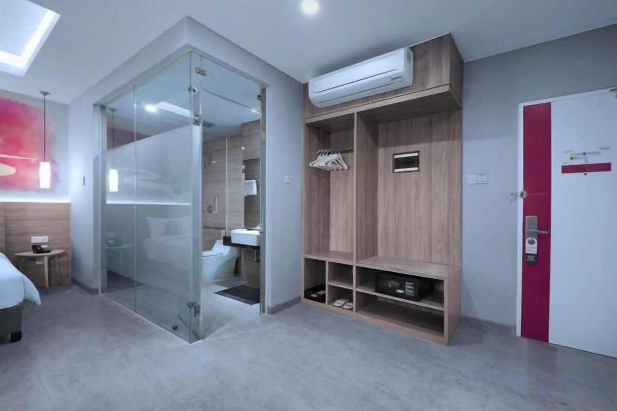 A modern hotel room with a glass partition, a wooden wardrobe, and a built-in air conditioning unit.