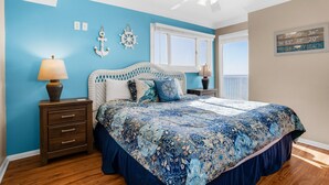 Une chambre à coucher aux couleurs bleu et blanc, avec un lit recouvert d’une couvre-lit à motifs, une table de chevet avec une lampe et une fenêtre donnant sur un paysage.