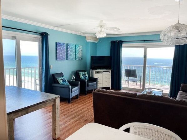 A living room with a view of the ocean, a ceiling fan, and a TV on a white cabinet.