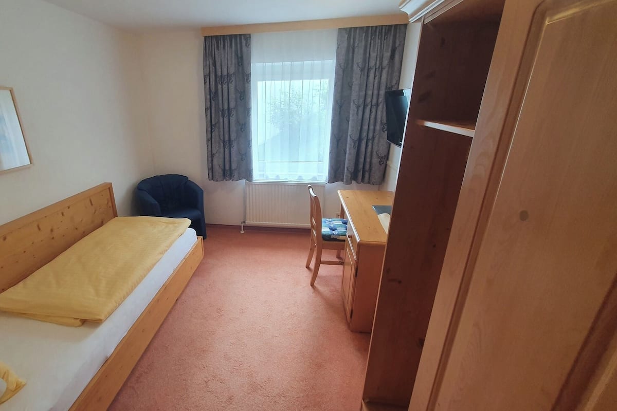 Una camera d'albergo piccola e singola, con un letto, una scrivania, una sedia, un termosifone e una finestra con tende.