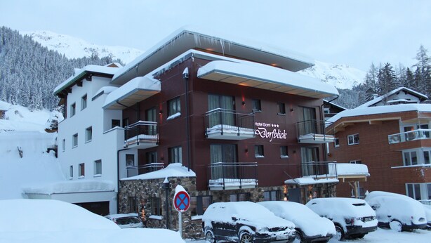 A hotel with a snow-covered exterior, multiple balconies, and a parking area with snow-covered cars. - Hotel Garni Dorfblick (Sankt Anton am Arlberg)