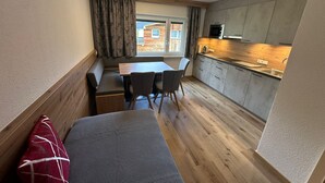 A compact room with a dining table, chairs, a bench, a kitchenette, and a window with curtains. - Hotel Garni Dorfblick (Sankt Anton am Arlberg)