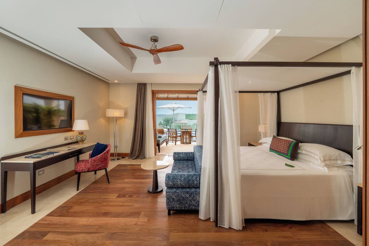A hotel room with a bed, a desk, a chair, a small table, and a view of the outdoors.