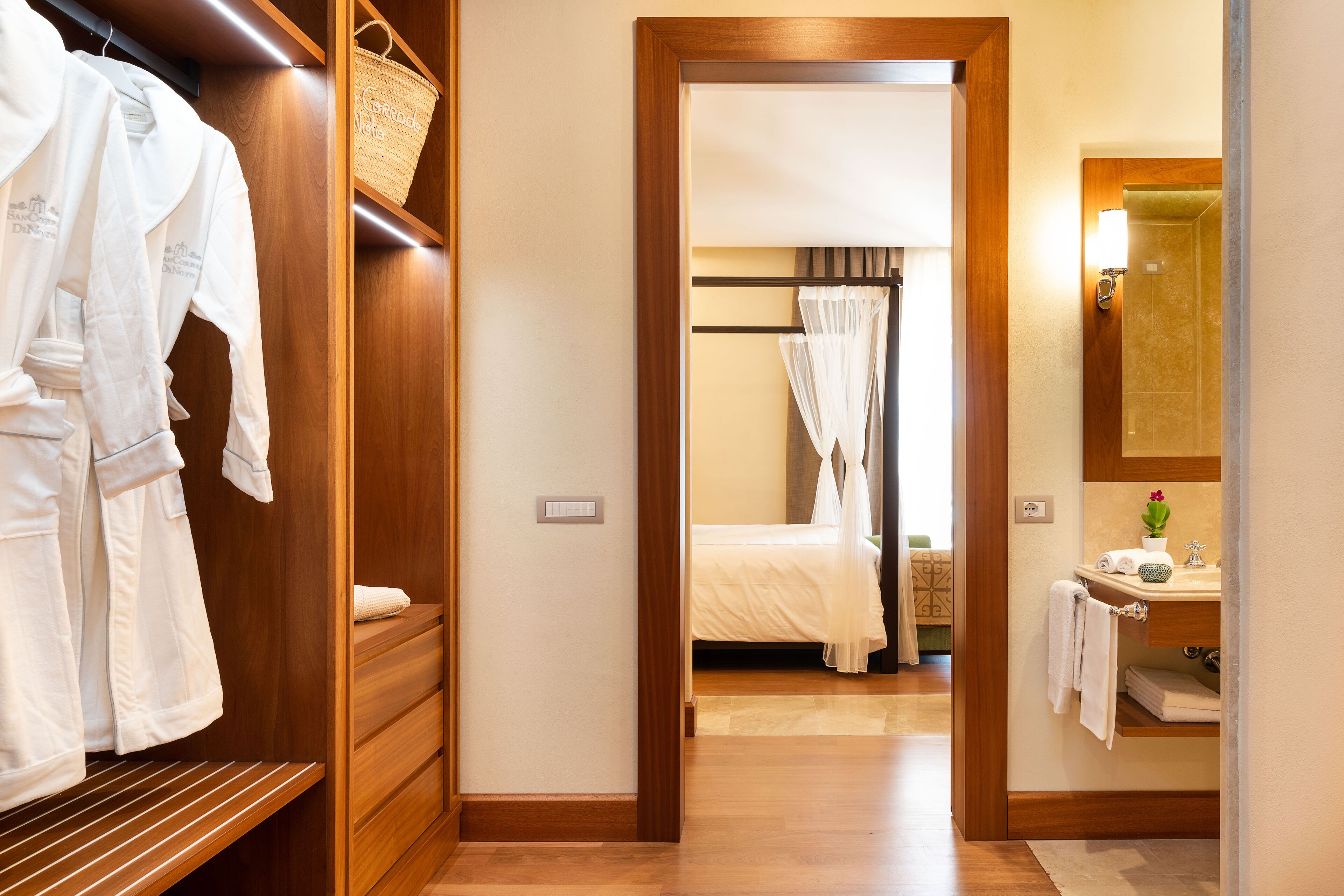 Habitación de hotel con armario de madera, cama con dosel, baño con espejo y lavamanos, y una mesita con una planta.