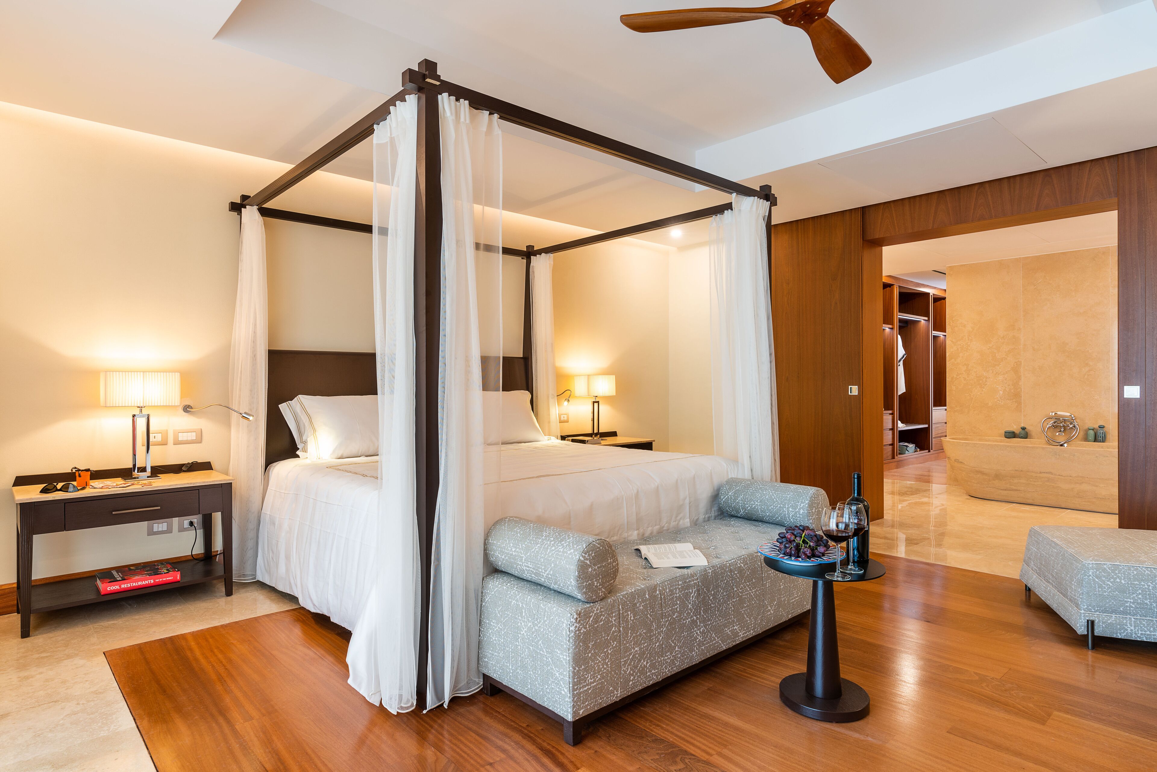 A four-poster canopy bed with a nightstand, a reading lamp, and a small table with a wine glass and grapes.