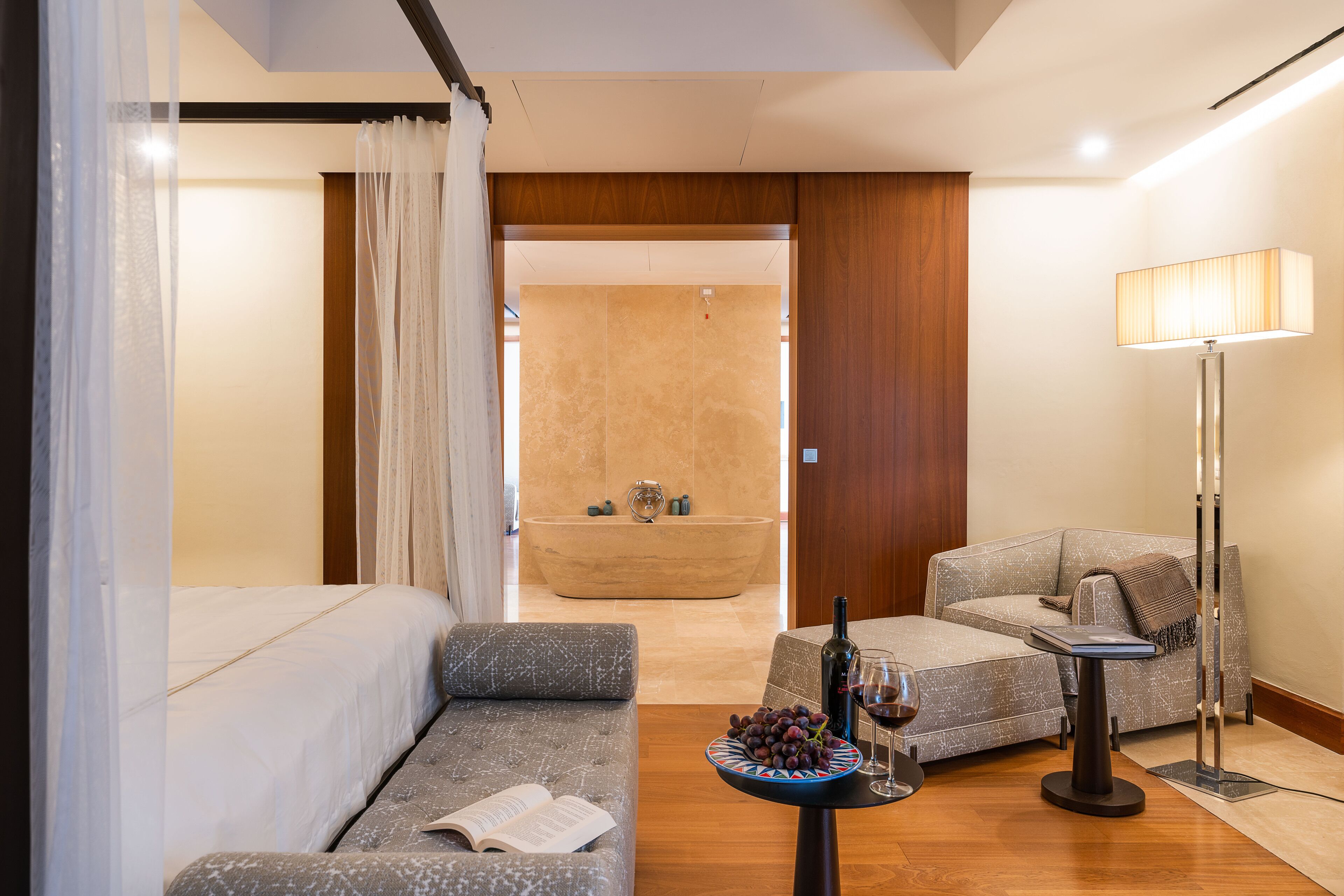 A modern hotel room with a sofa, a small table with a bottle and grapes, a floor lamp, and a view of a bathroom with a bathtub.