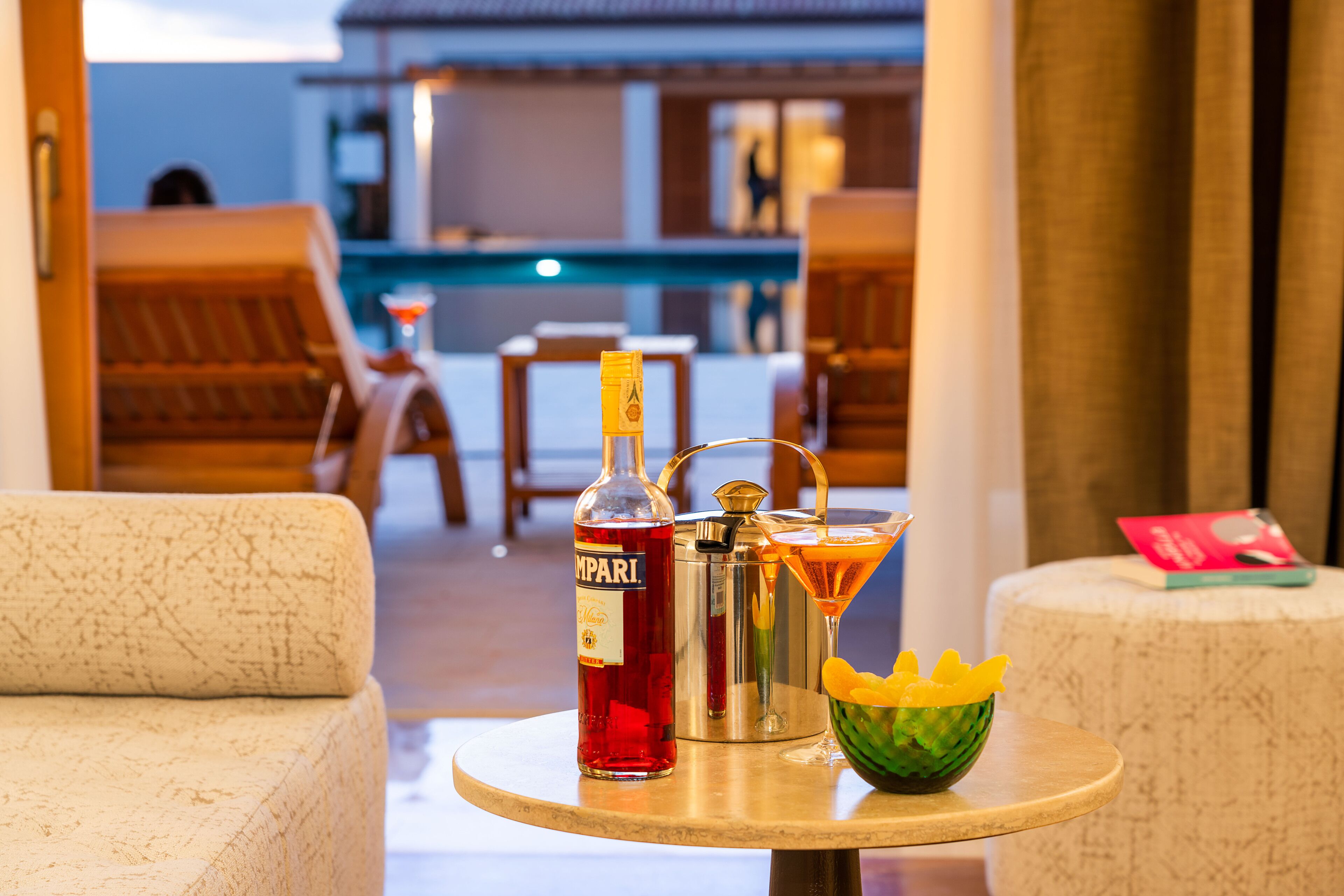 Ein runder Tisch mit einer Spirituosenflasche, einem Cocktail-Shaker und einem Cocktail mit einer Garnitur, vor einer Poollandschaft mit Liegestühlen.