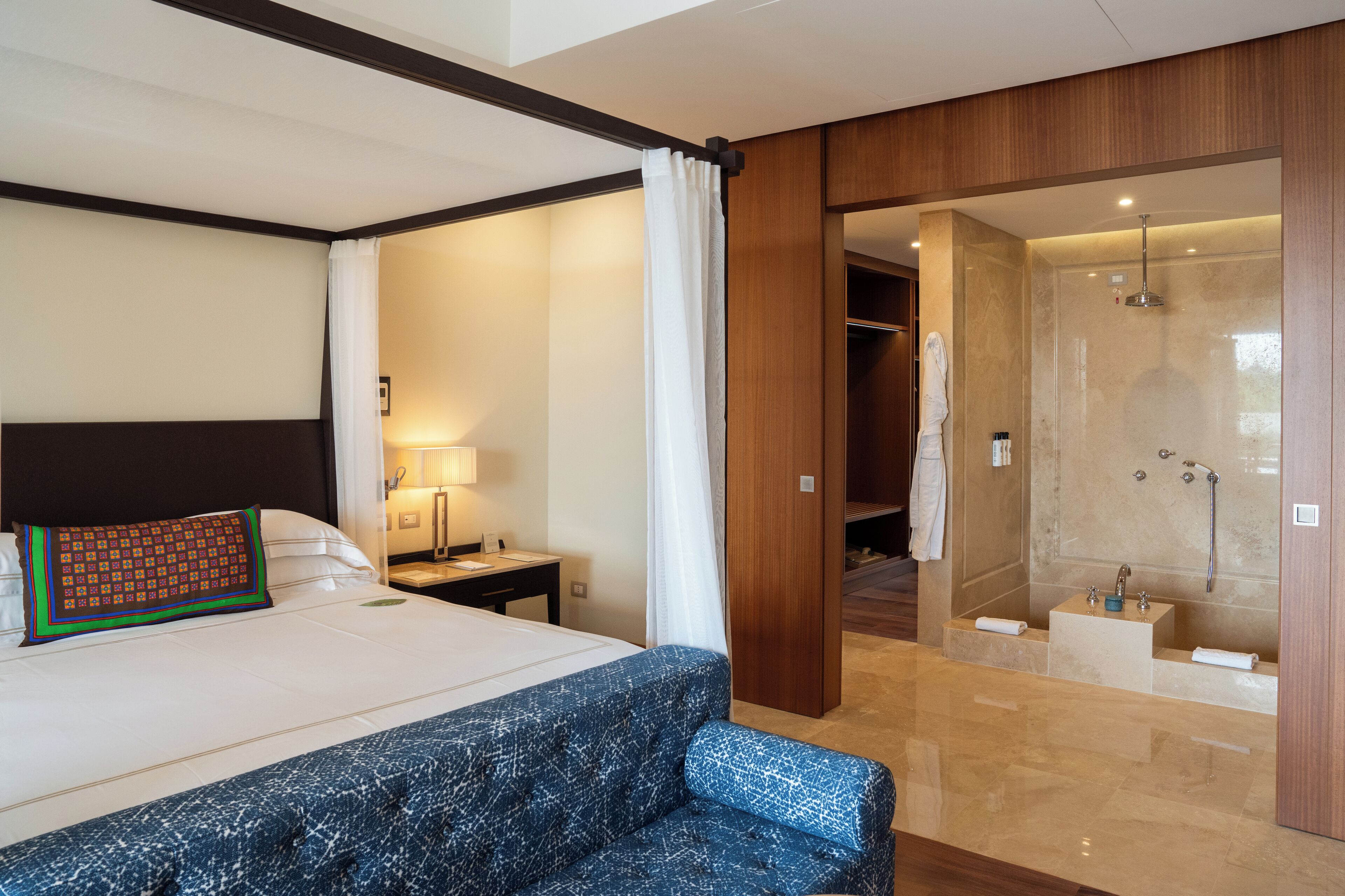 Habitación de hotel con una cama grande, una mesita de noche, un escritorio, zona de ducha y baño con lavabo y espejo.