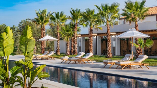 A poolside area with lounge chairs and umbrellas, surrounded by palm trees and a building with a tiled roof.