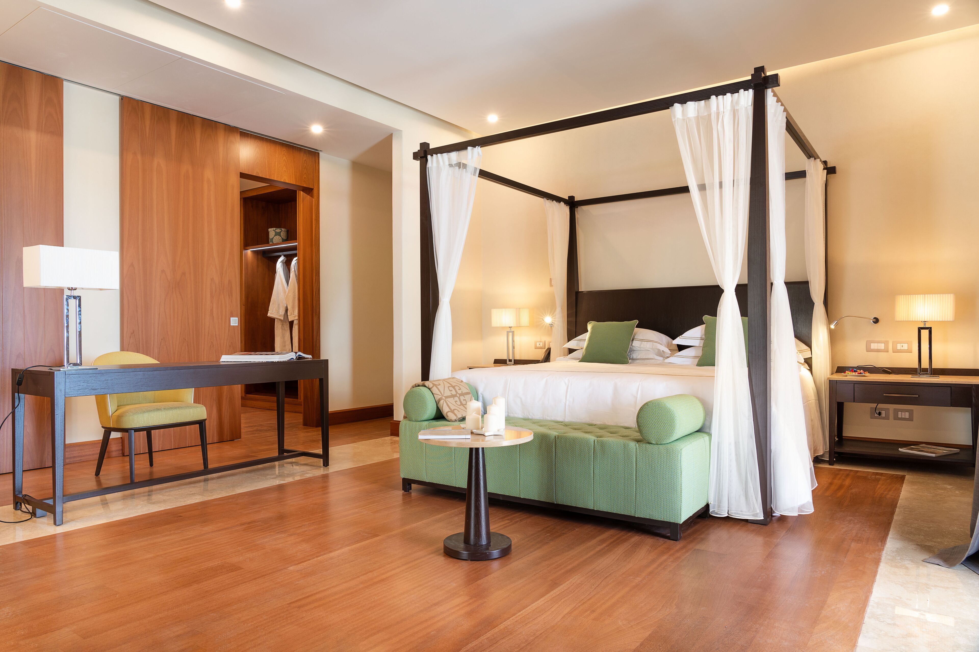Ein Hotelzimmer mit Himmelbett, Schreibtisch mit Stuhl, einem kleinen Tisch und einem grünen Sofa.