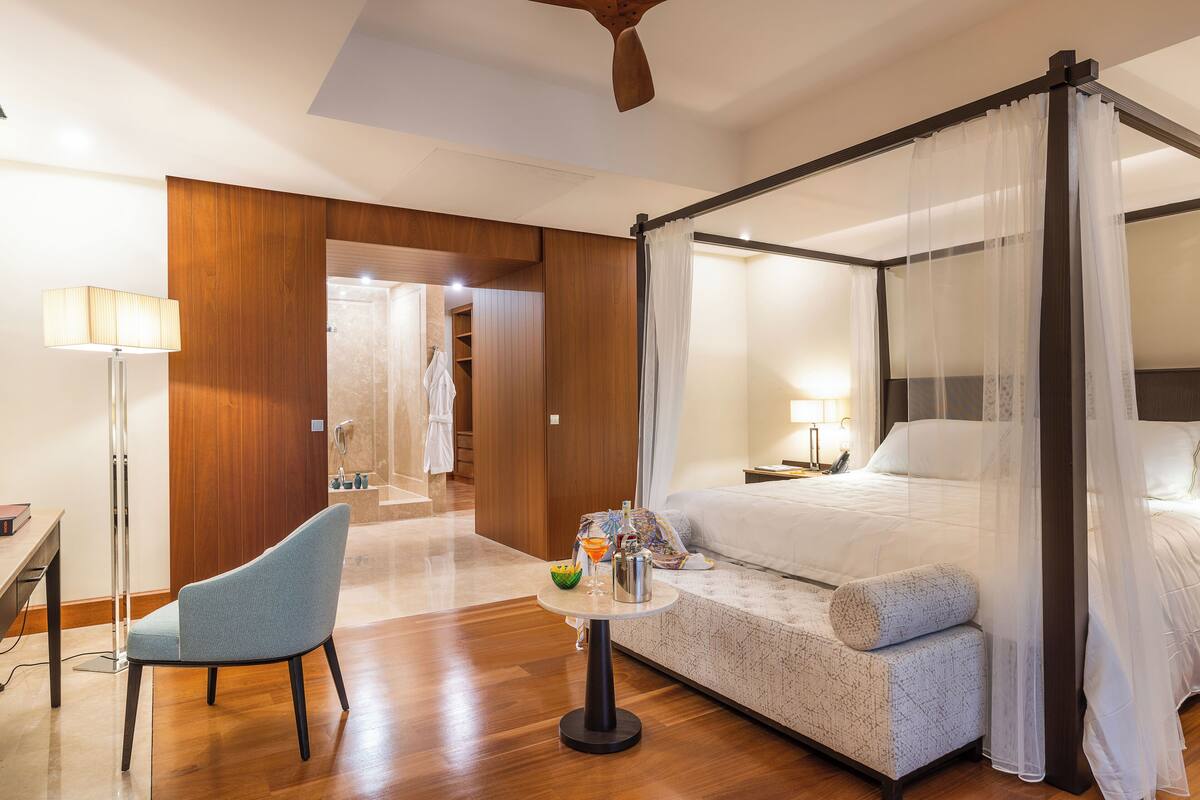 A modern hotel room with a four-poster bed, a sitting area with a table, and a separate desk.