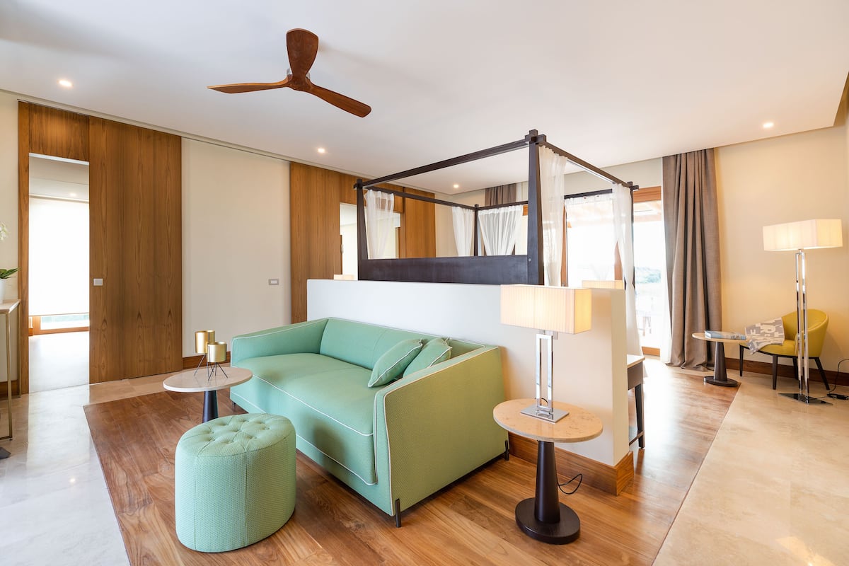 A modern hotel room with a green sofa, a round ottoman, a ceiling fan, a bed with a canopy, and a small table with a lamp.
