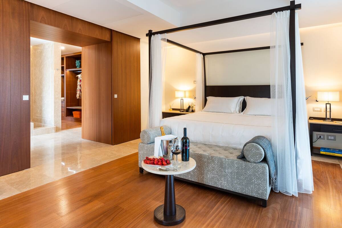 A four-poster bed with a canopy, a small round table with a bottle and glasses, and a sofa with cushions.