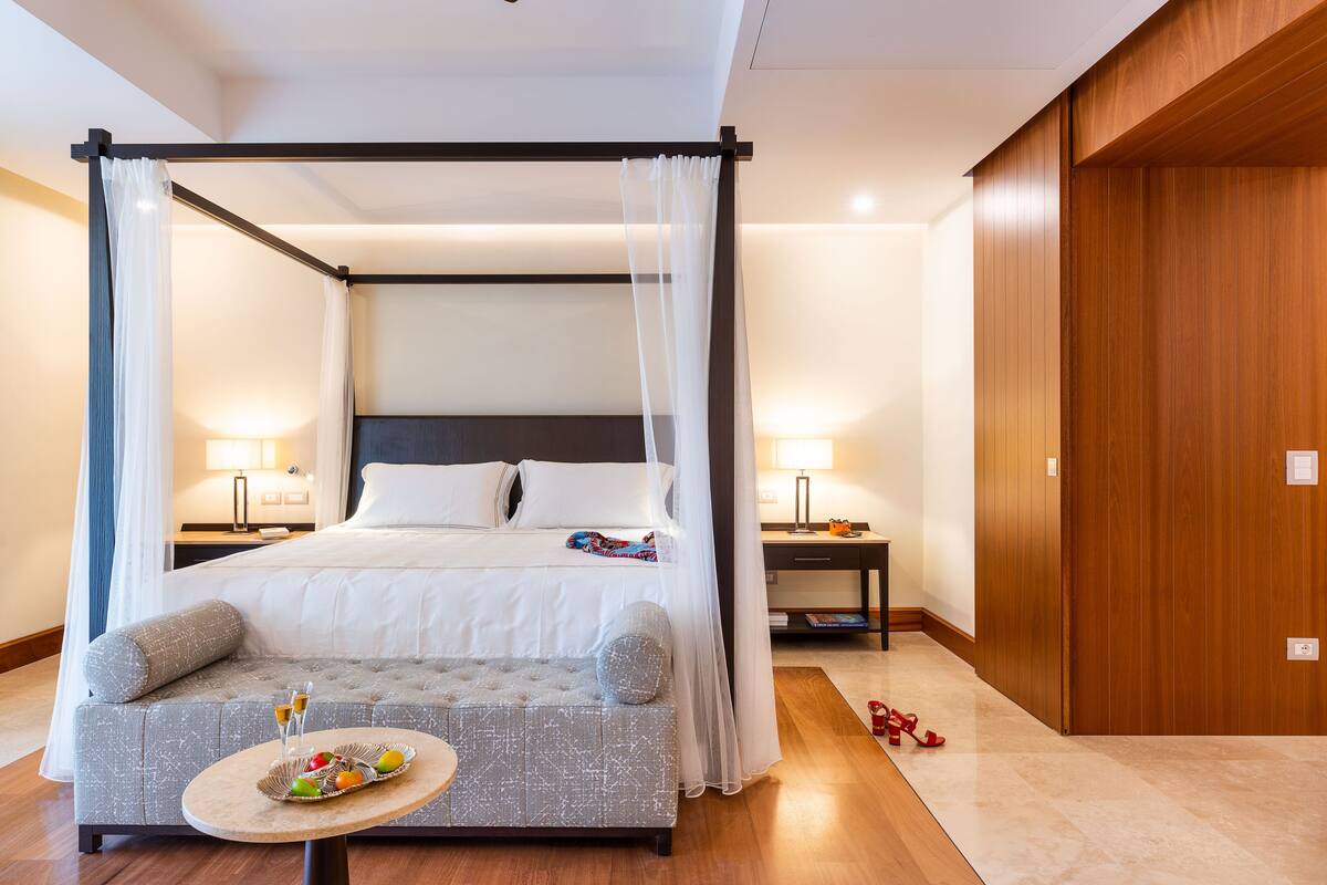 A hotel room with a four-poster bed, a sofa, a small table with fruit and drinks, and a wooden wardrobe.