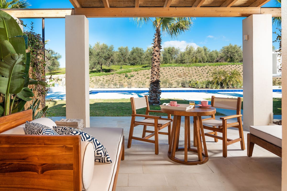 A covered outdoor seating area with a pool view, wooden furniture, and a view of the surrounding landscape.