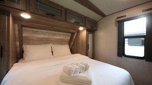 Cama bem arrumada com roupa de cama branca e toalhas dobradas, cabeceira de madeira e janela com persianas.
