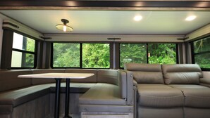 Interior compacto de uma caravana, com área de estar acolchoada, mesa pequena e grandes janelas com vista para o verde.