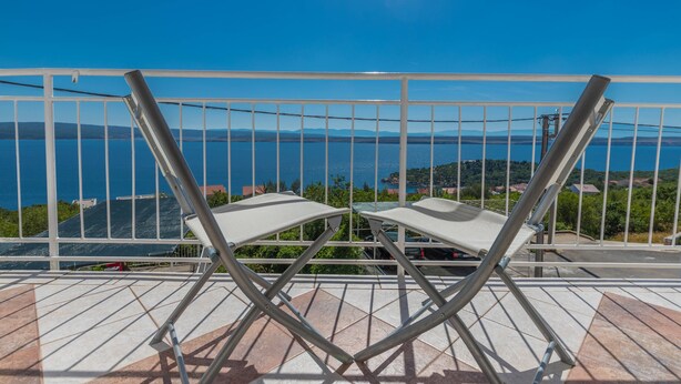 Een balkon met twee witte klapstoelen, een metalen hekwerk en uitzicht op een waterpartij en verre heuvels.