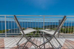 A balcony with two white folding chairs, a metal railing, and a view of a body of water and distant hills.
