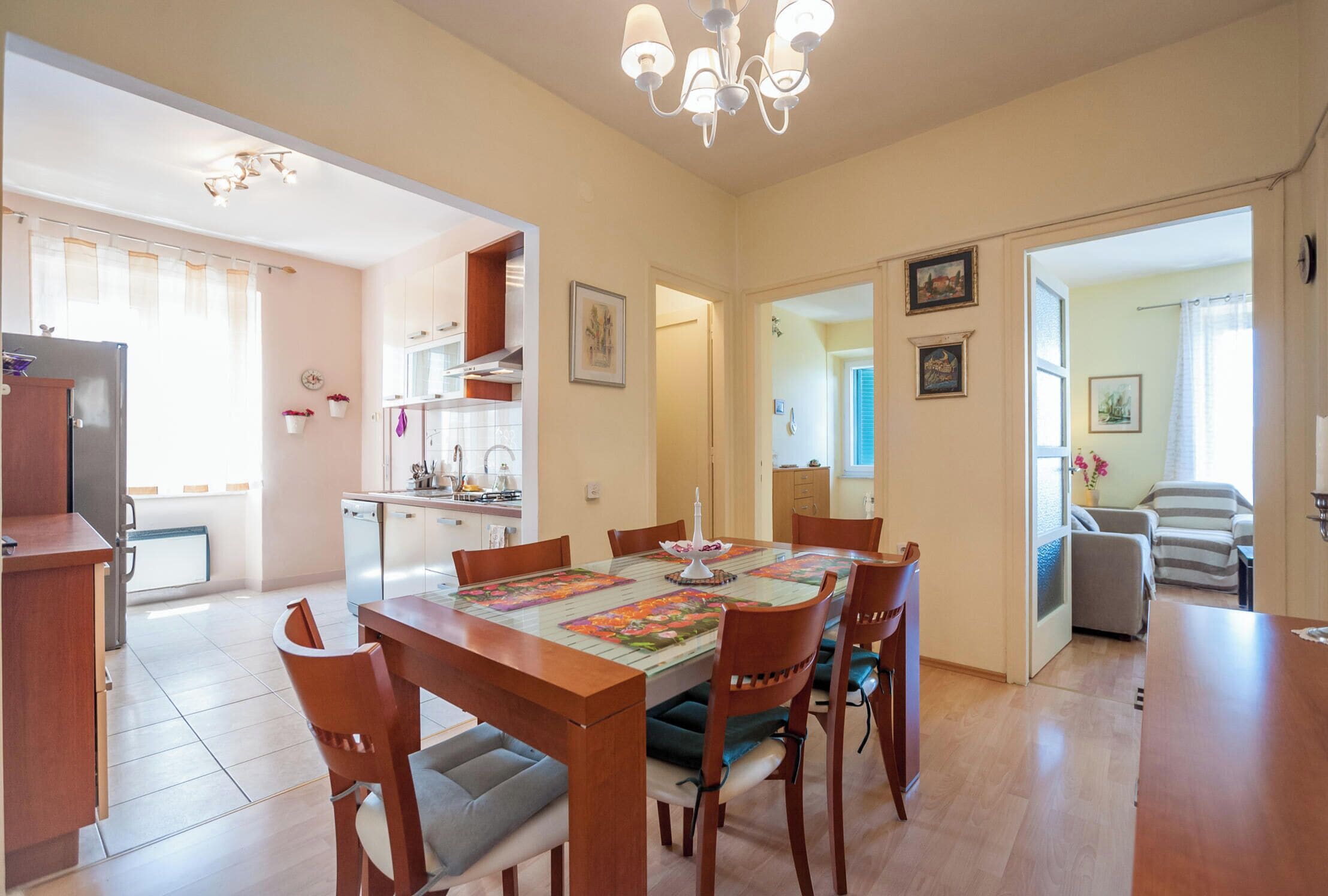 A dining area with a wooden table and chairs, a chandelier, and a kitchen in the background.