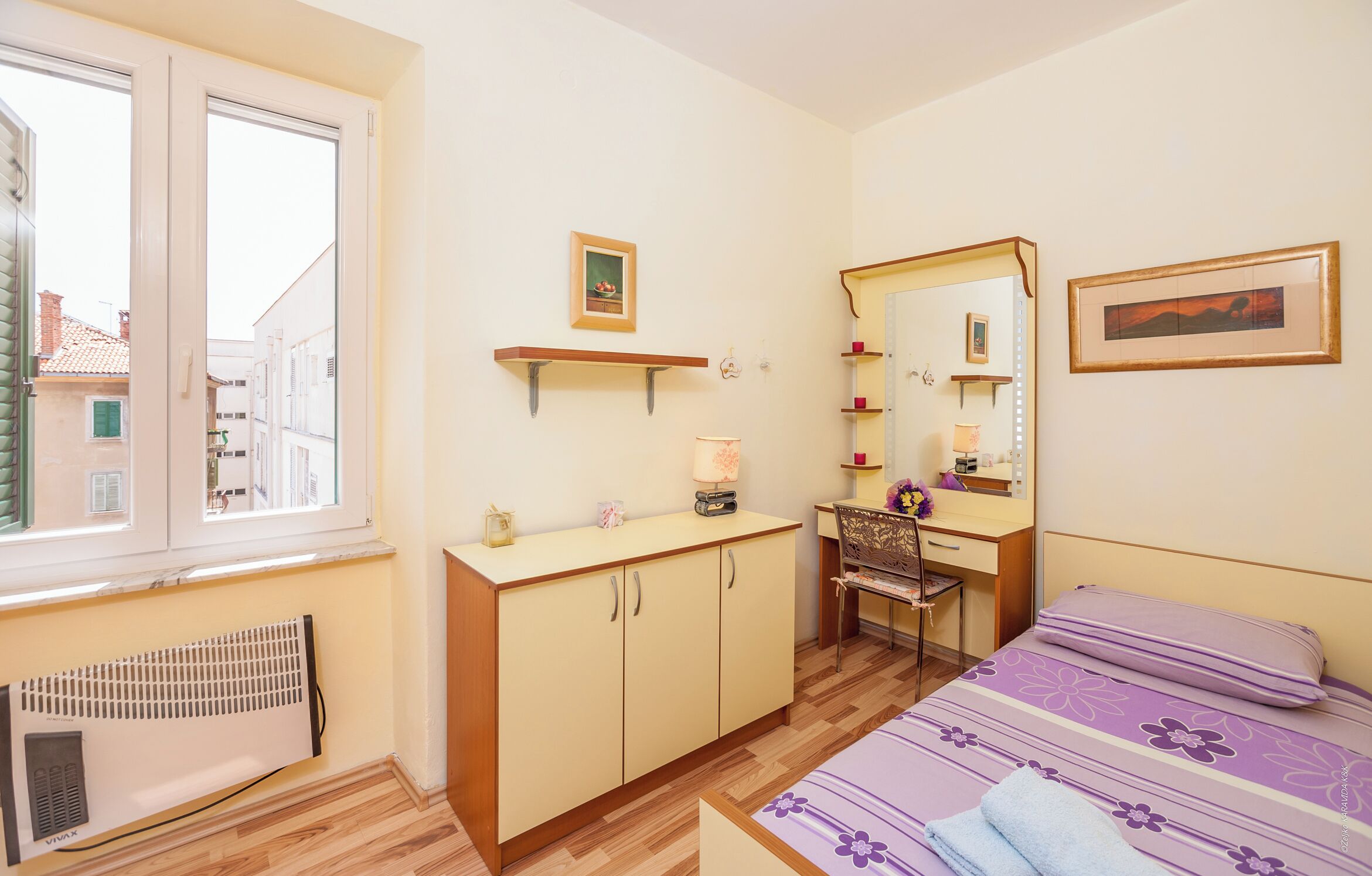 A bedroom with a bed, a desk, a chair, a radiator, and a window.