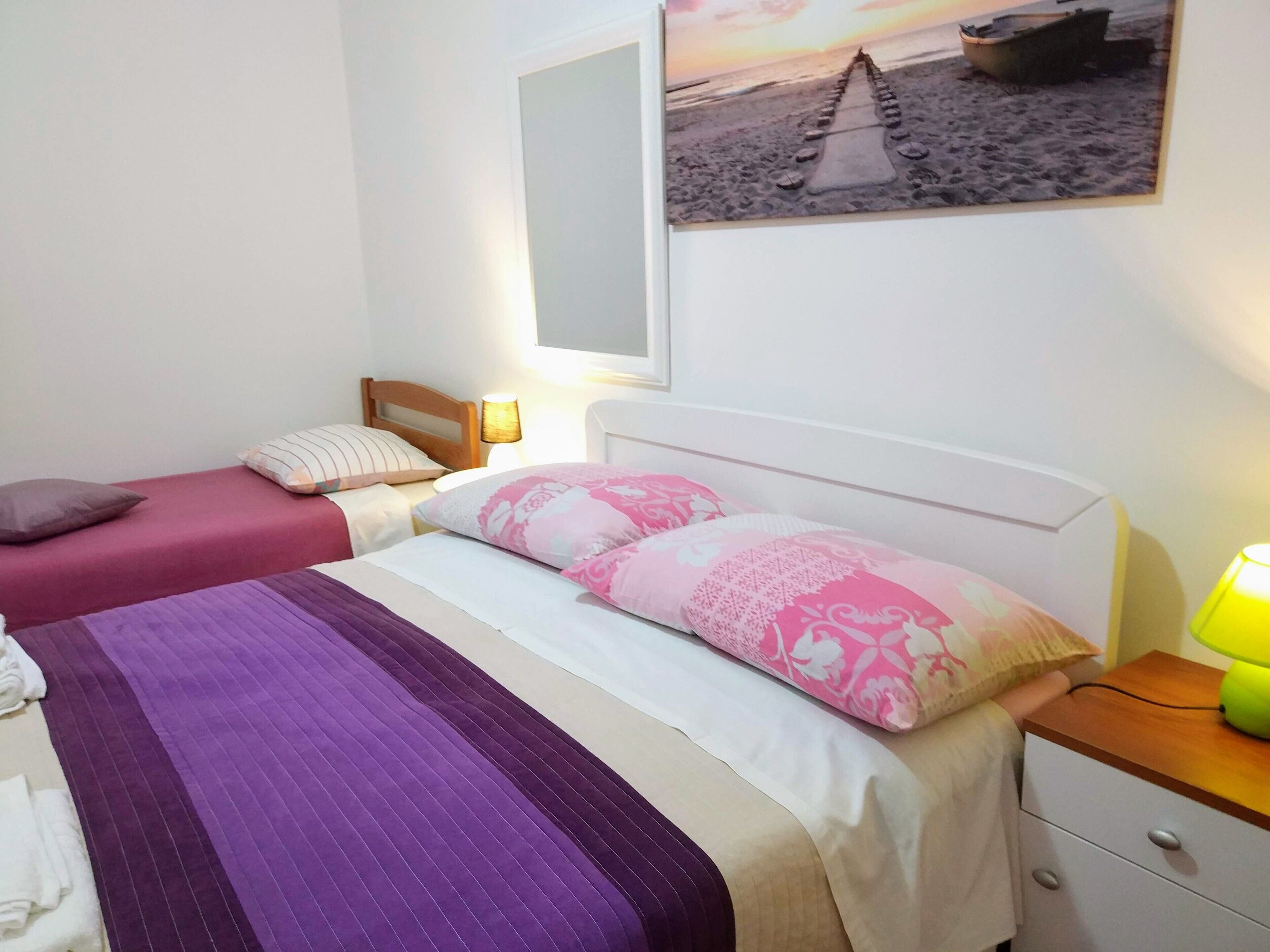A double bed with a purple and white bedspread, a single bed with a red bedspread, a nightstand with a lamp, and a wall-mounted picture of a beach.