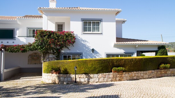 A white building with a tiled roof, stone wall, and a well-maintained hedge.